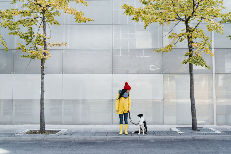 Woman wearing yellow raincoat looking at dog while standing on footpath during autumn