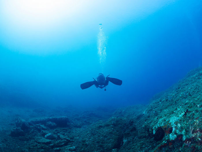 Unrecognizable professional diver in wetsuits and flippers swimming near rough stony formation with uneven surface in deep blue sea with clear water