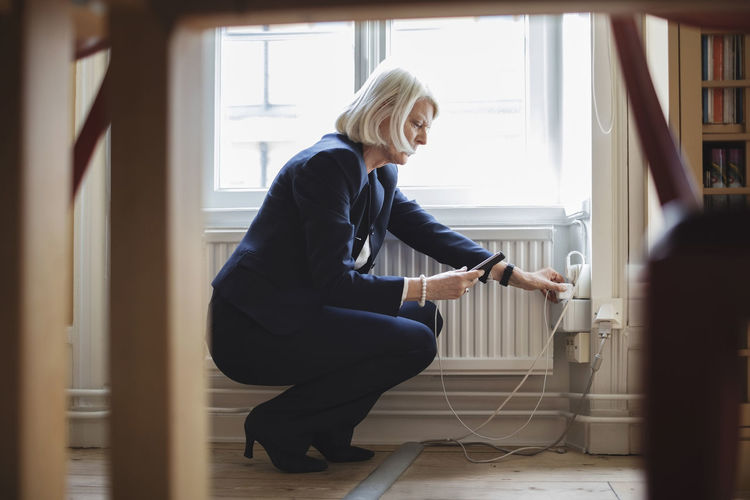 Senior woman crouching while charging mobile phone by radiator with furniture in foreground at library