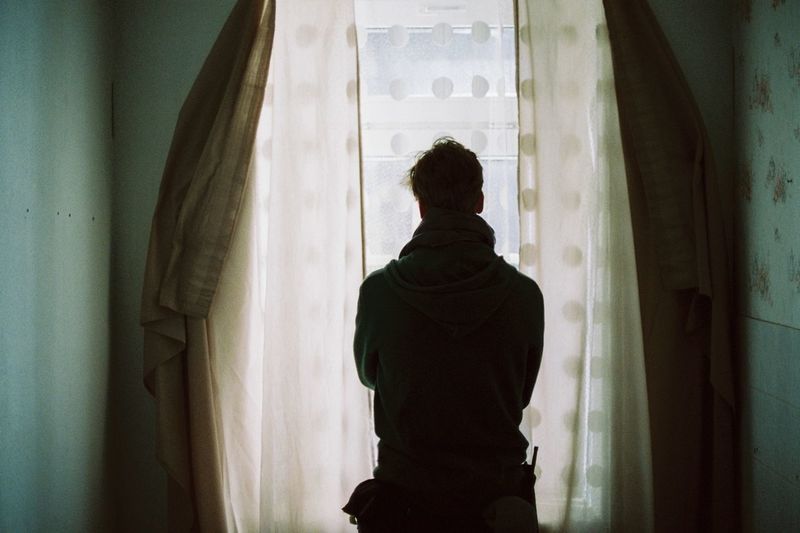 Rear view of man looking through curtained window