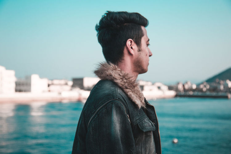 Side view of man wearing jacket while standing by sea against clear sky in city