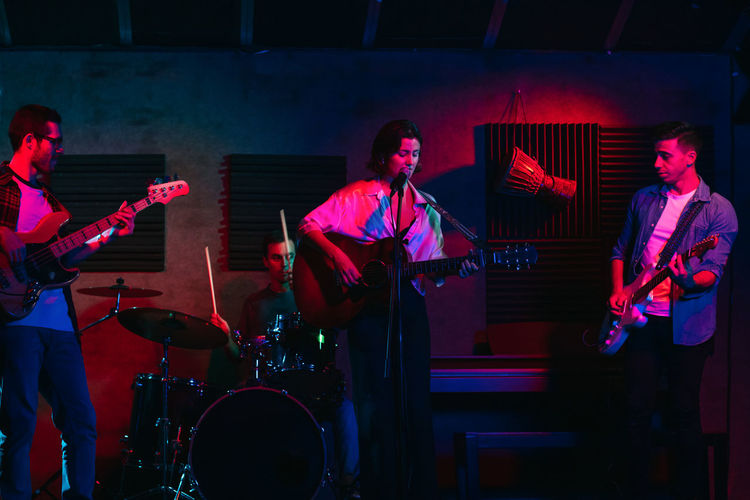 Group of people in casual clothes playing guitars and drums while woman singing and performing song in club with neon lights