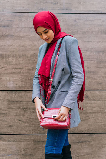 Side view of young content arab female in hijab and trendy clothes standing near building in city and looking down