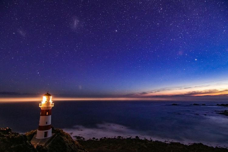 Illuminated lighthouse by sea against sky at night
