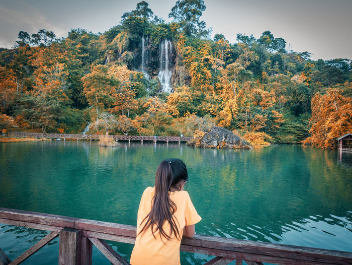 Rear view of woman standing by lake against waterfall