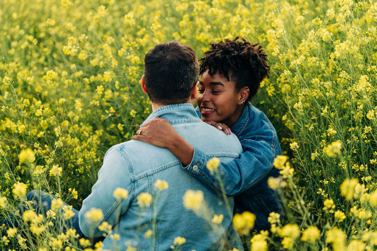 Loving young multiracial couple in casual clothes looking at each other while sitting in lush blooming meadow during romantic date on sunny day