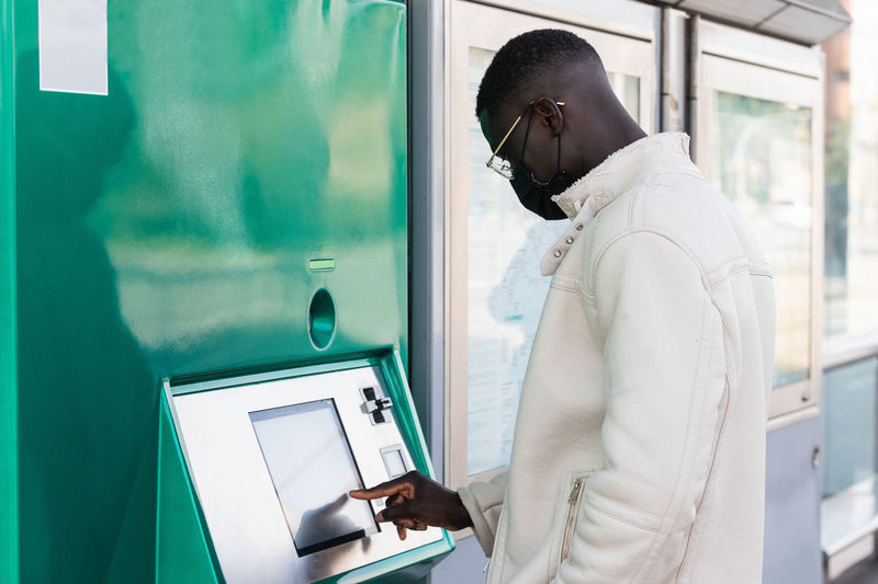 Side view of ethnic male entrepreneur in cloth face mask using automated teller machine in city
