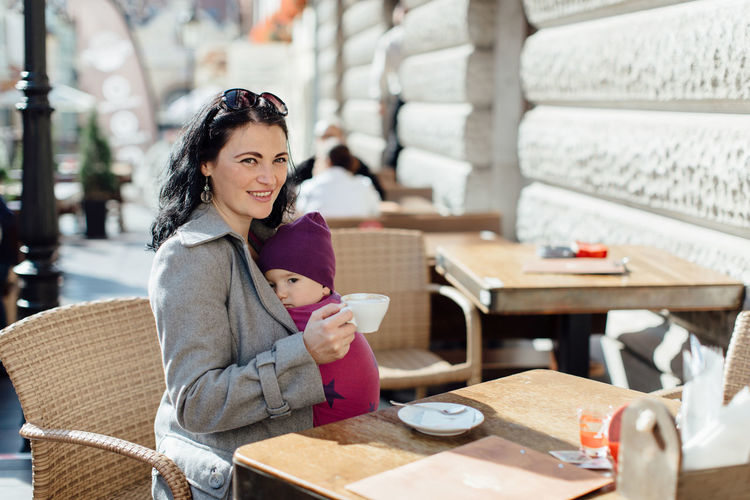 Portrait of woman carrying son in scarf on chair at sidewalk cafe