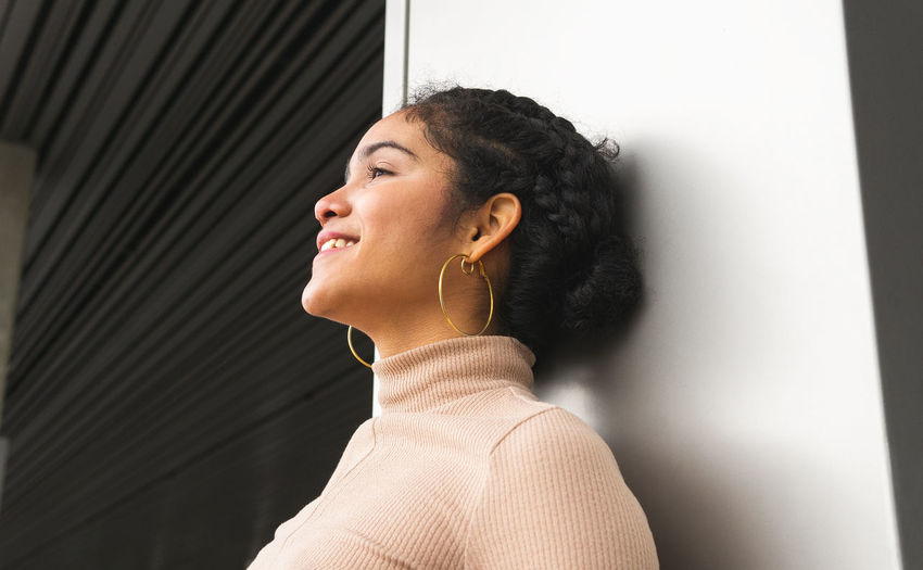 From below side view of happy colombian female in turtleneck standing near gray wall and looking away with interest