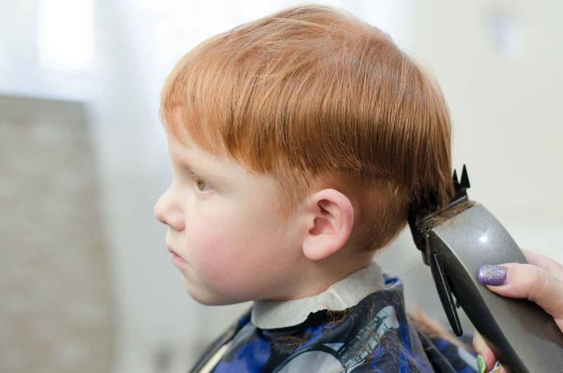 Red-haired boy at the barbershop. the hands of the hairdresser cut the boy with a trimmer