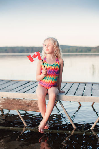 Happy caucasian girl sitting on pier by lake waving canadian flag. smiling child holding canada flag 
