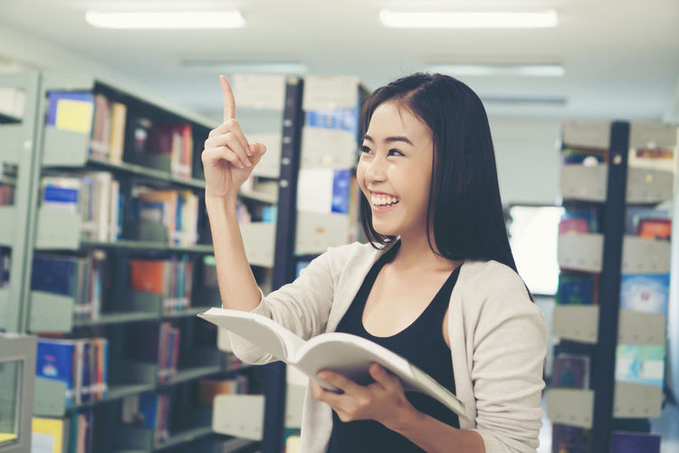 Smiling young woman pointing while holding book in library