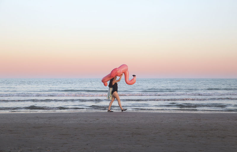 Young woman walking with inflatable pink flamingo on the beach at sunset