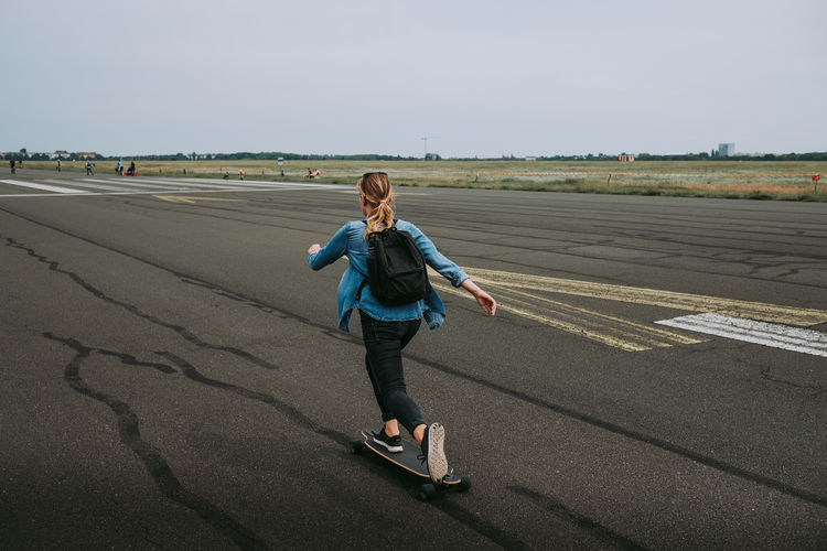 Rear view of woman with backpack skateboarding on road against sky