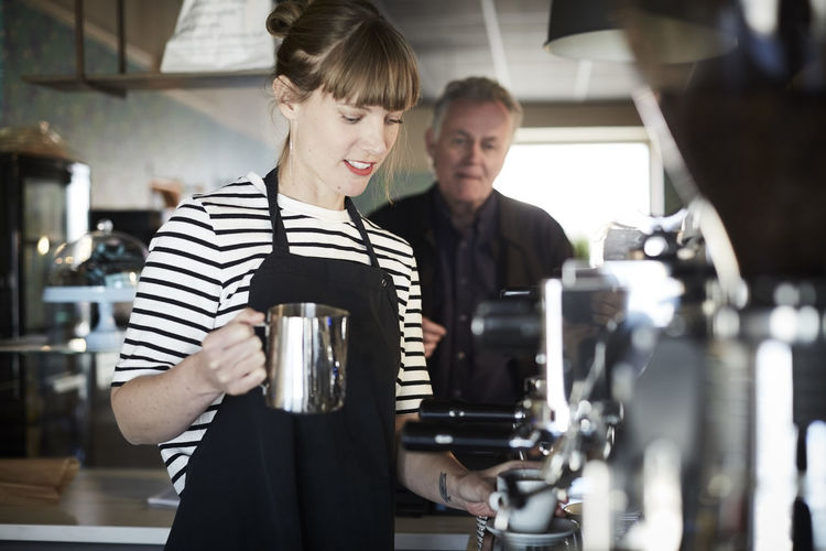 Female barista holding pitcher while making coffee with customer standing in background at cafe