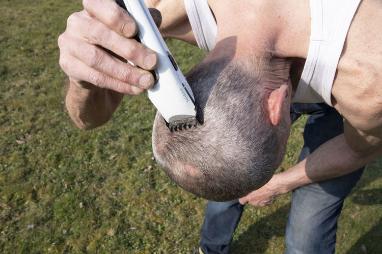 A gray-haired middle-aged man shaves his hair with a clipper in a garden