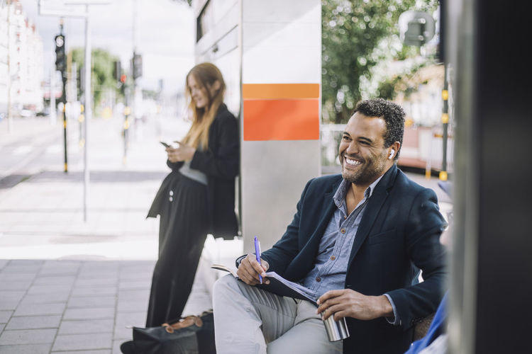 Happy male freelancer wearing wireless in-ear headphones sitting with diary against businesswoman at bus stop