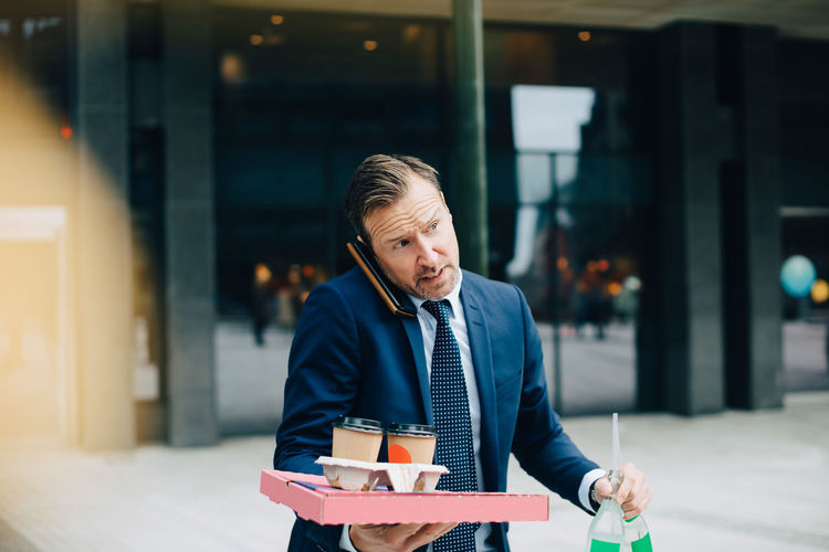 Mature businessman talking on mobile phone while carrying food and drinks in city