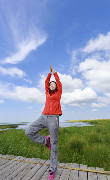 Young woman doing yoga on boardwalk by grassy field against sky