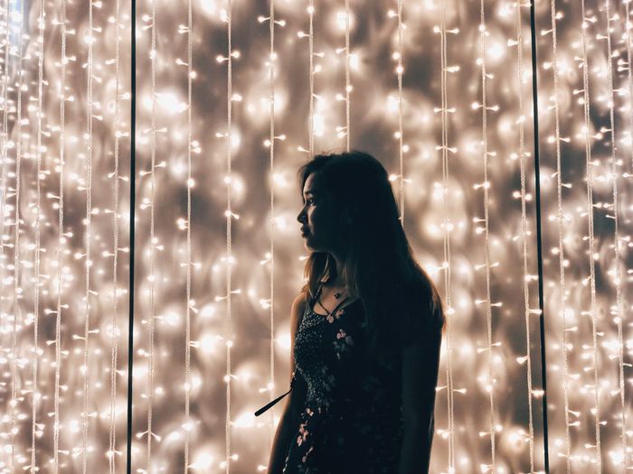Young woman looking away while standing against illuminated string lights at night