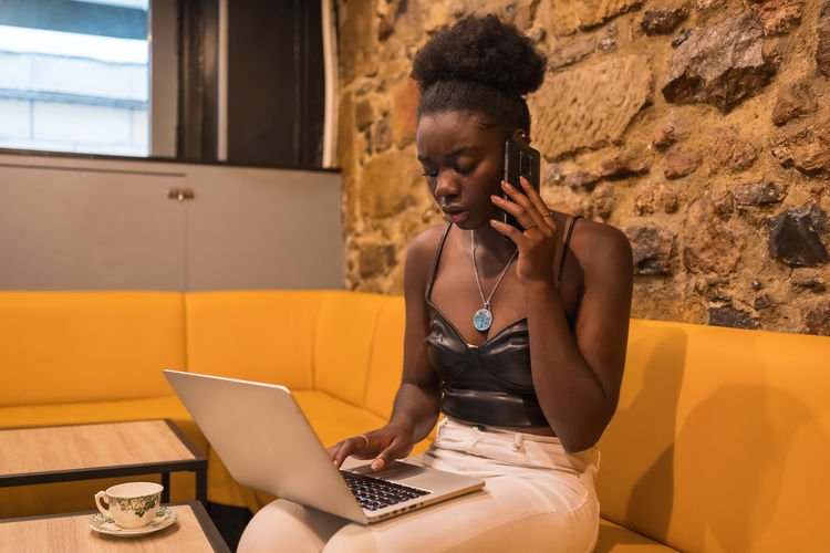 Side view of busy african american female freelancer speaking on mobile phone while sitting at table with laptop in cafe and working on project