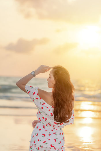 Rear view of woman with arms outstretched standing at beach against sky during sunset