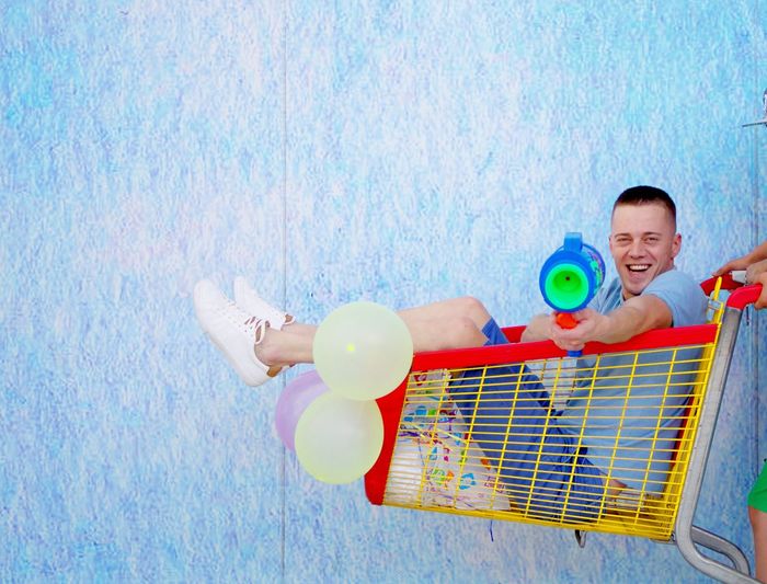 Portrait of smiling man holding squirt gun while sitting in shopping cart by wall