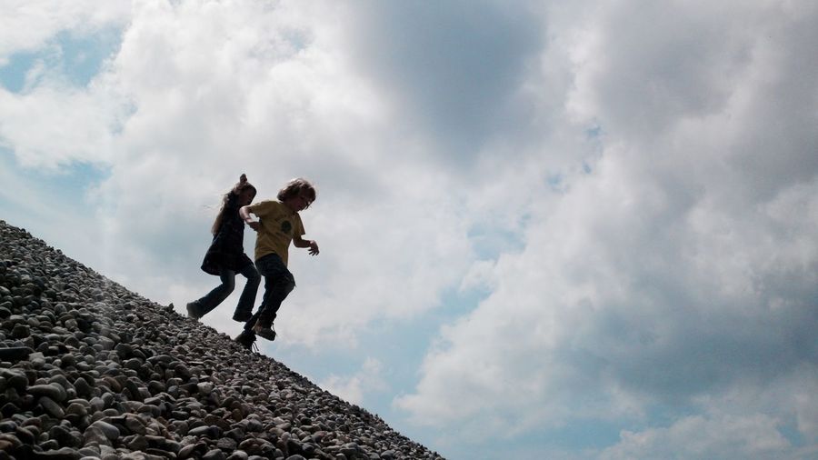 Siblings walking on heap of stones at construction site against cloudy sky
