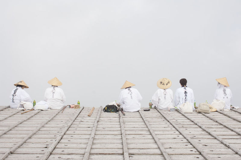 Rear view of people wearing asian style conical hats sitting in row against sky