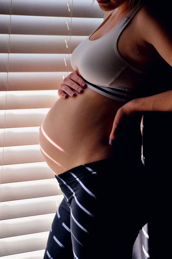 Midsection of pregnant woman standing by window blinds