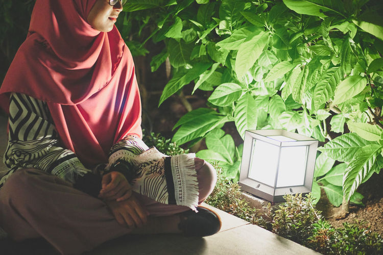 Smiling young woman sitting on walkway by illuminated lamp and plants