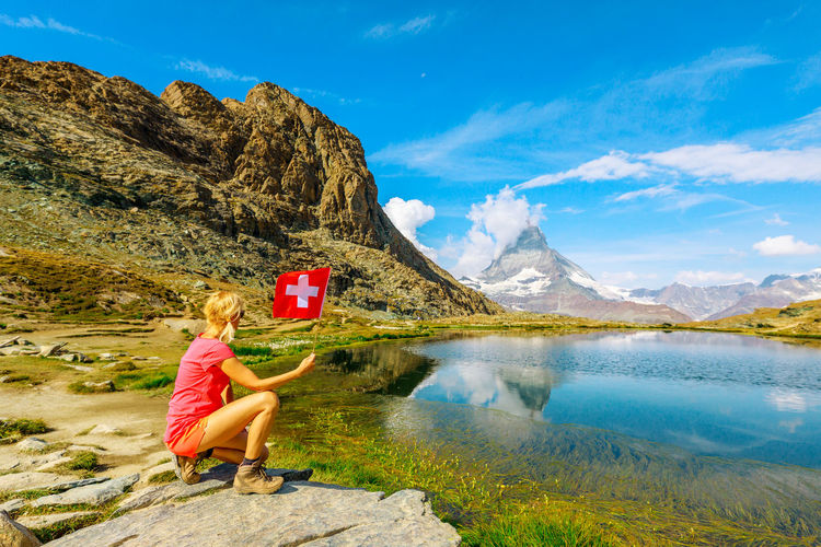 Rear view of woman crouching on rock while holding flag by lake against sky