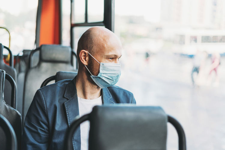 Adult bald bearded man in medical face mask looking out the window in bus