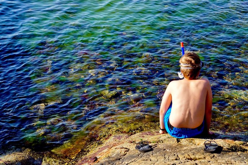 Rear view of shirtless boy sitting on rock by lake