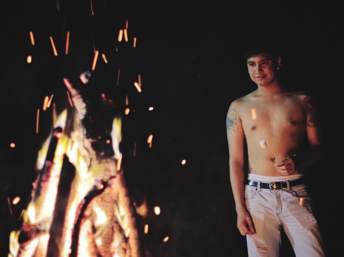 Shirtless man holding marshmallow while standing by bonfire