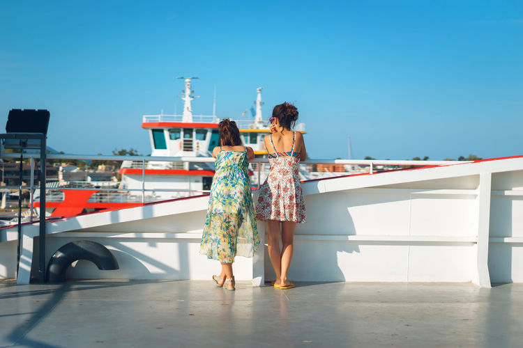 Rear view of women standing by railing on boat against blue sky