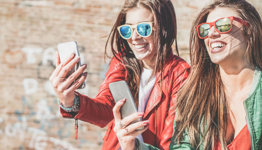 Happy friends taking self portrait on mobile phones during sunny day