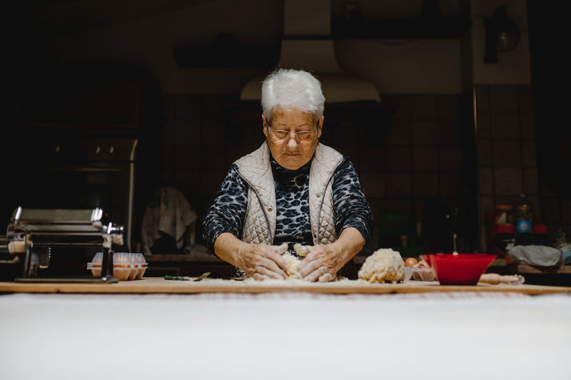 Senior female standing at table in kitchen and kneading dough while preparing ingredients for cooking appetizing tortellini