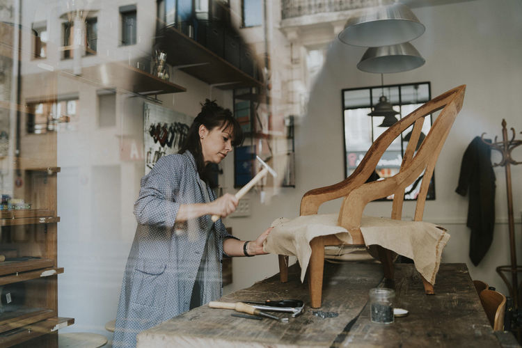 Female upholsterer making wooden chair on table in workshop seen through window