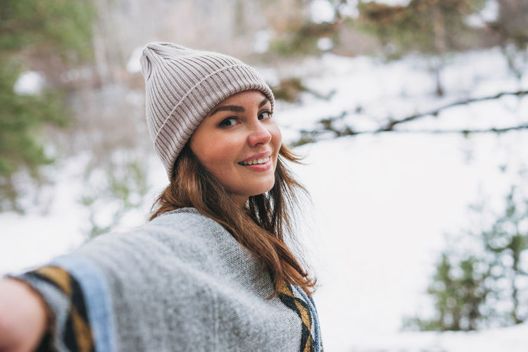 Young brunette beautiful woman in hat and grey poncho walking in the winter forest