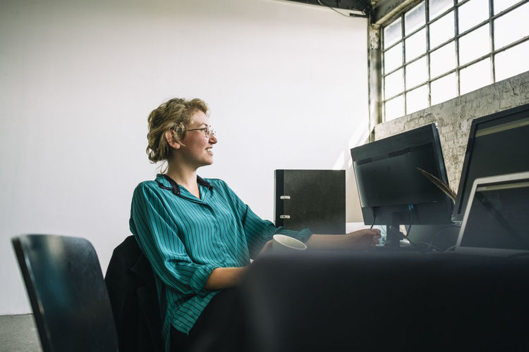 Thoughtful young businesswoman smiling and looking away while sitting at desk in creative office
