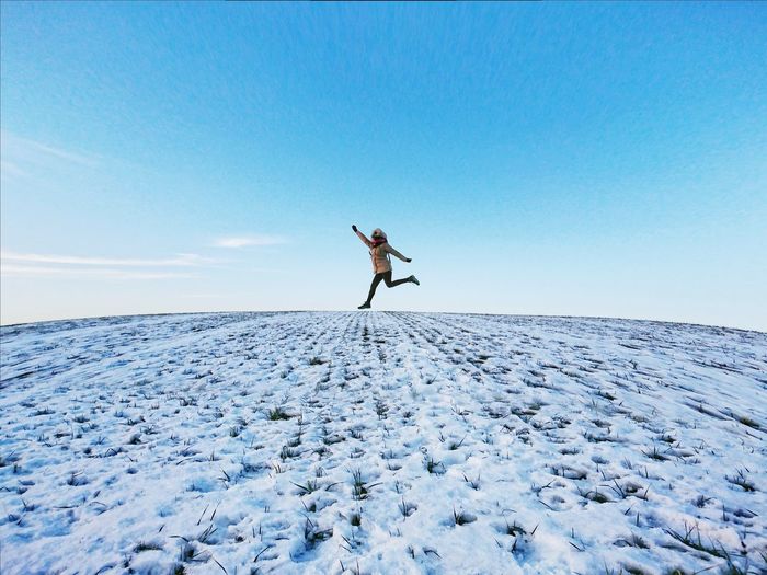 Mid adult woman jumping on snow covered land against blue sky