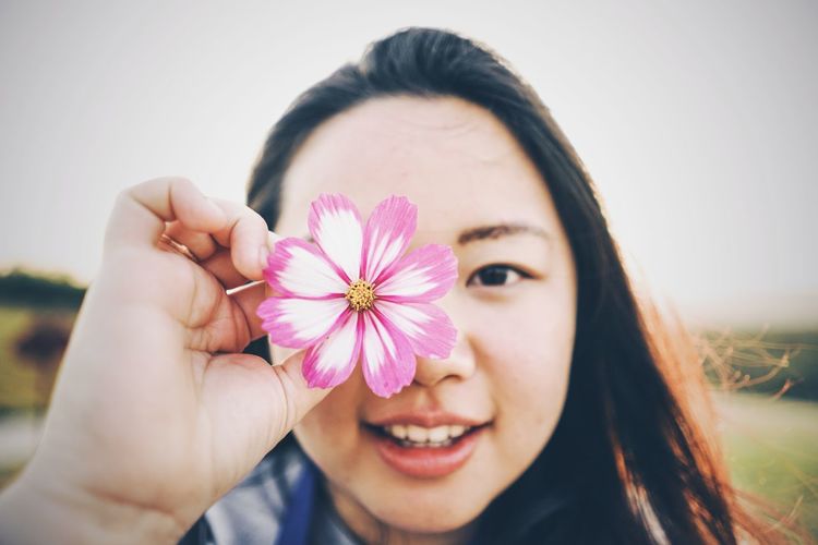 Close-up portrait of young woman holding pink flower against sky