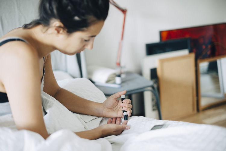 Woman checking blood sugar level while sitting on bed in bedroom