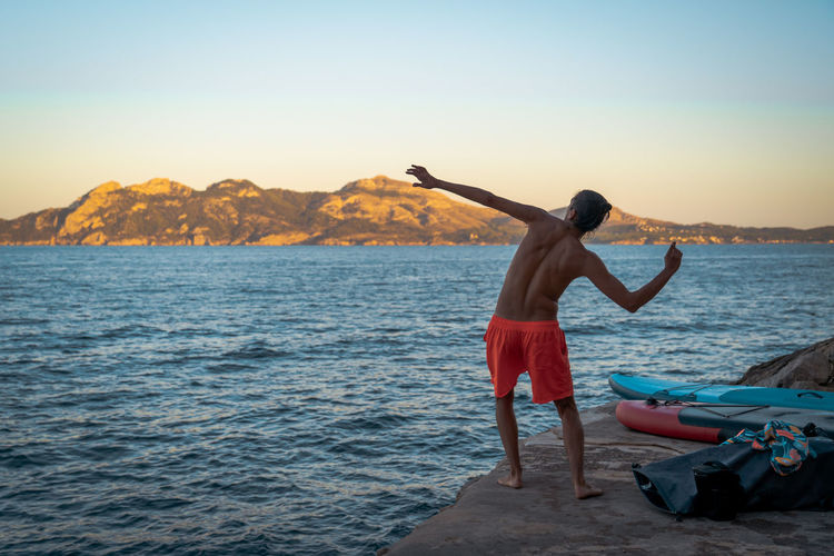 Rear view of shirtless man throwing rock to sea