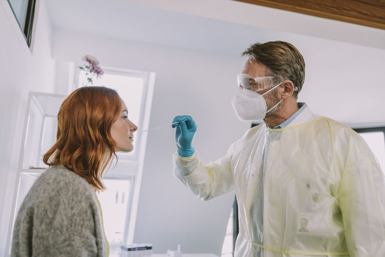 Doctor in protective suit and face mask checking patient while standing in examination room