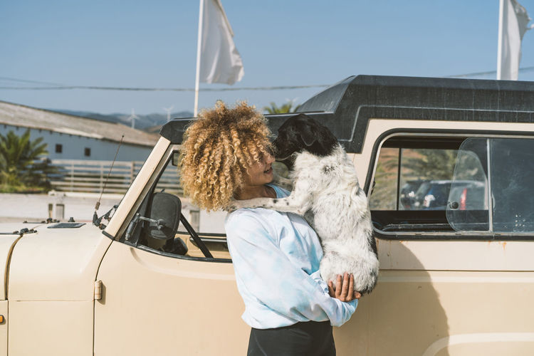 Young blond afro woman carrying dog while standing by old off-road vehicle on sunny day
