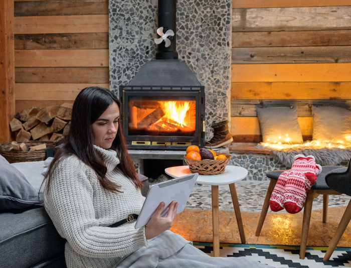 Young woman in warm sweater sitting by fireplace, using tablet computer.