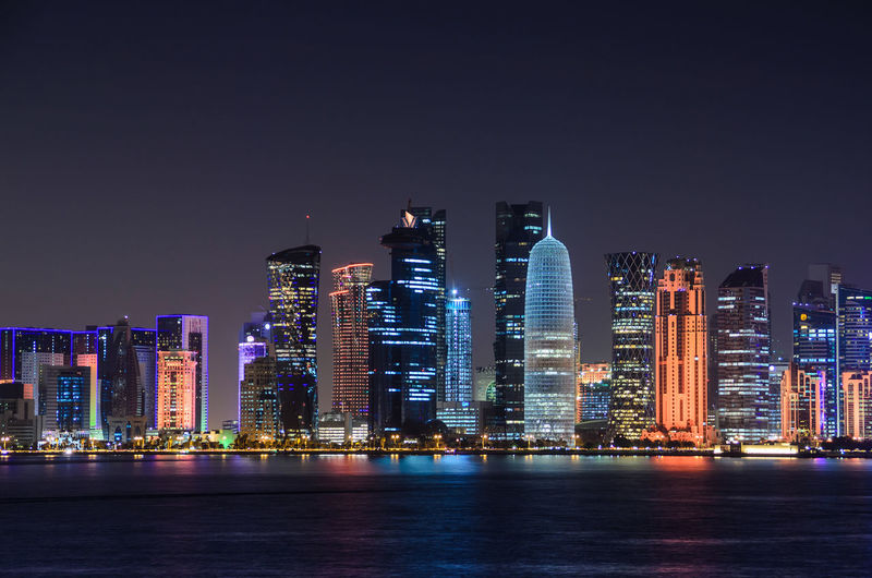 Illuminated doha tower and skyscrapers by river in city against clear sky at night