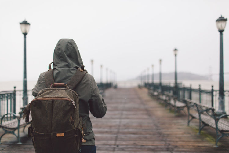 Rear view of young woman with backpack walking on pier against sky